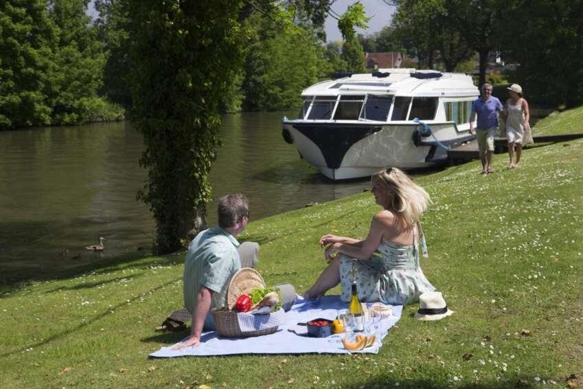 Viajar en barco | Danfluvial Pareja disfrutando de un picnic en la orilla del río junto a su barco en una escapada de turismo fluvial con Danfluvial.