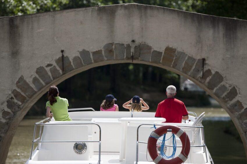 Turismo fluvial | Danfluvial Pasajeros disfrutando de la experiencia del turismo fluvial a bordo de un barco de recreo que cruza bajo un puente de piedra.