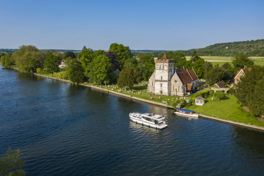 Barco fluvial navegando por el río Támesis junto a una iglesia histórica en la campiña inglesa.
