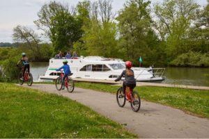 Dos personas paseando en bici por el sendero de tierra junto a un barco fluvial que navega por el canal.